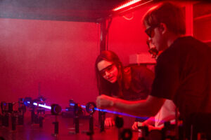 Three students wearing laser safety glasses work together in a photonics laboratory, aligning a bright blue laser beam through a series of optical lenses and mirrors on an optical table illuminated by red lighting.